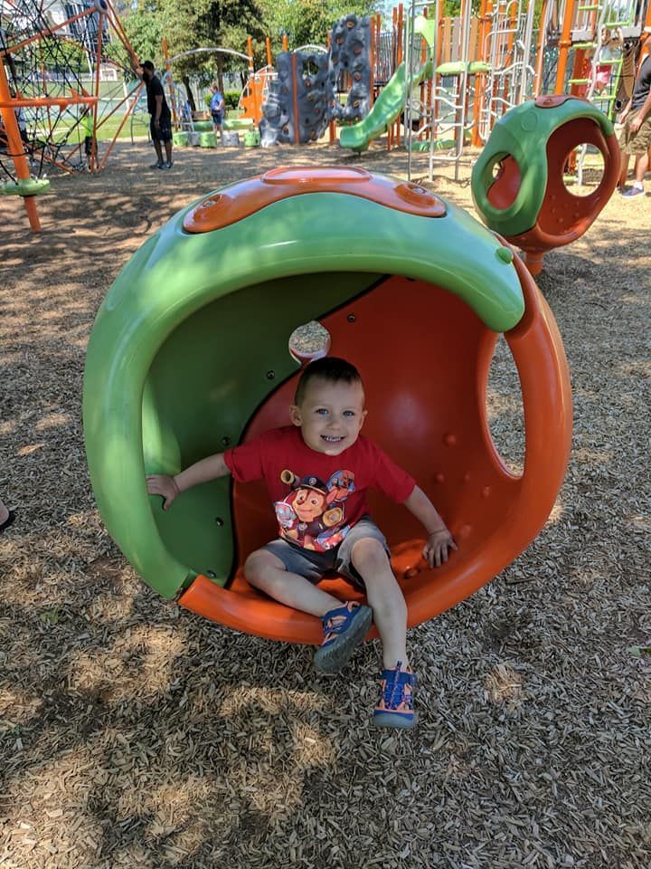 Boy sits inside a green and orange playground sphere, smiling. Playground in the background.