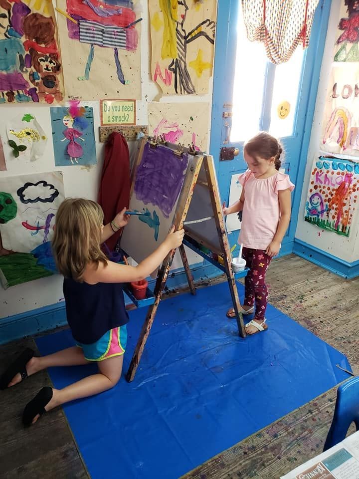 Two young girls painting at an easel in a colorful art room; one kneels to paint, the other watches.