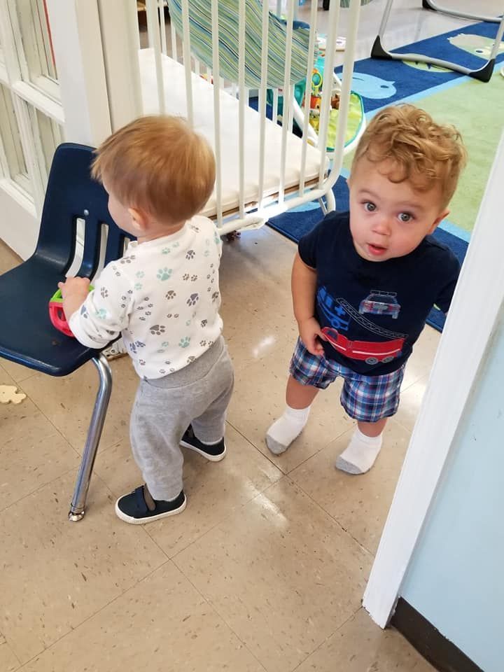 Two toddlers in a room. One stands, other leans on chair. One wears blue shirt and shorts, other wears white shirt and grey pants.