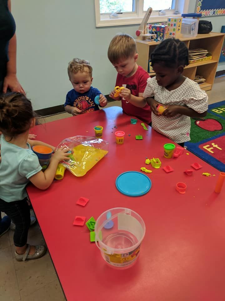 Children at a red table playing with Play-Doh. A teacher watches.