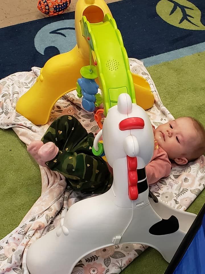 Baby laying on a blanket near a white toy and a yellow slide. Green and blue rug in background.
