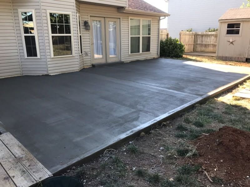 Newly poured concrete patio next to a house with a small wooden deck and a shed in the background.