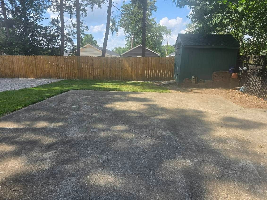 A backyard with a concrete patio, wooden fence, grass, and a green shed on a sunny day.
