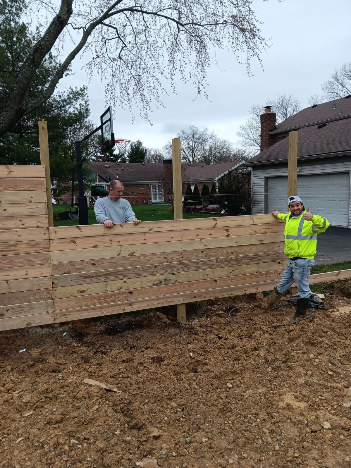Two men constructing a wooden fence in a residential yard; one gives a thumbs-up.
