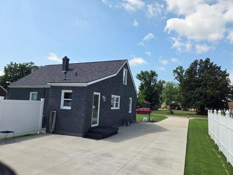 Gray brick house with dark roof, white trim, driveway, and white fence under blue sky with clouds.