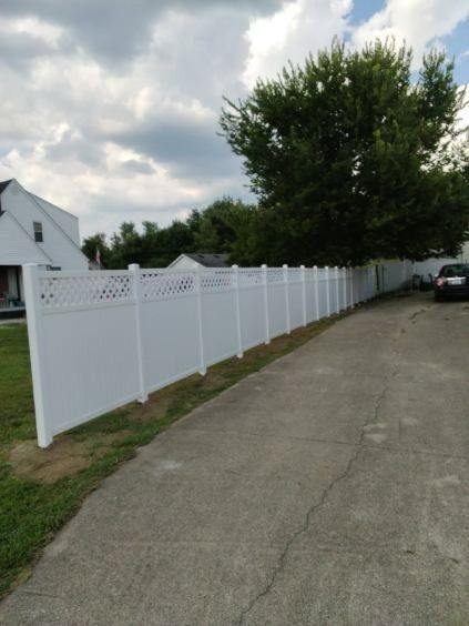 White vinyl fence with lattice top, along a concrete driveway, near a house and tree under cloudy sky.