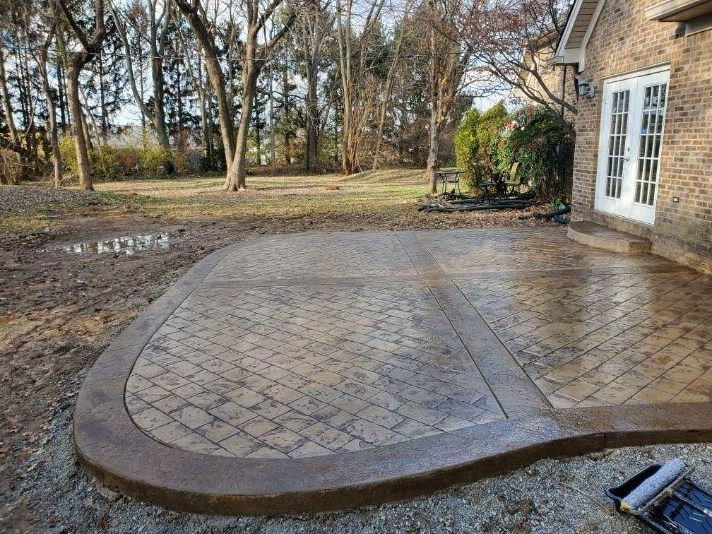 Concrete patio with brick pattern edging next to a brick house and grassy yard.