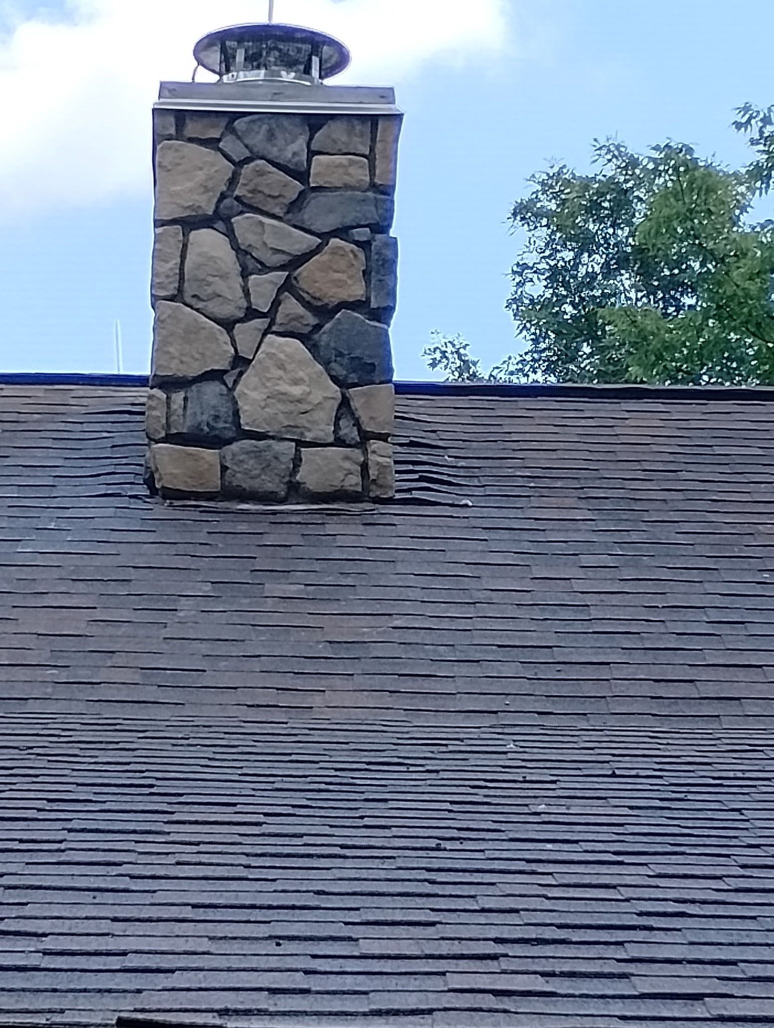 Stone chimney on a dark shingle roof with a cap, against a blue sky with tree foliage.