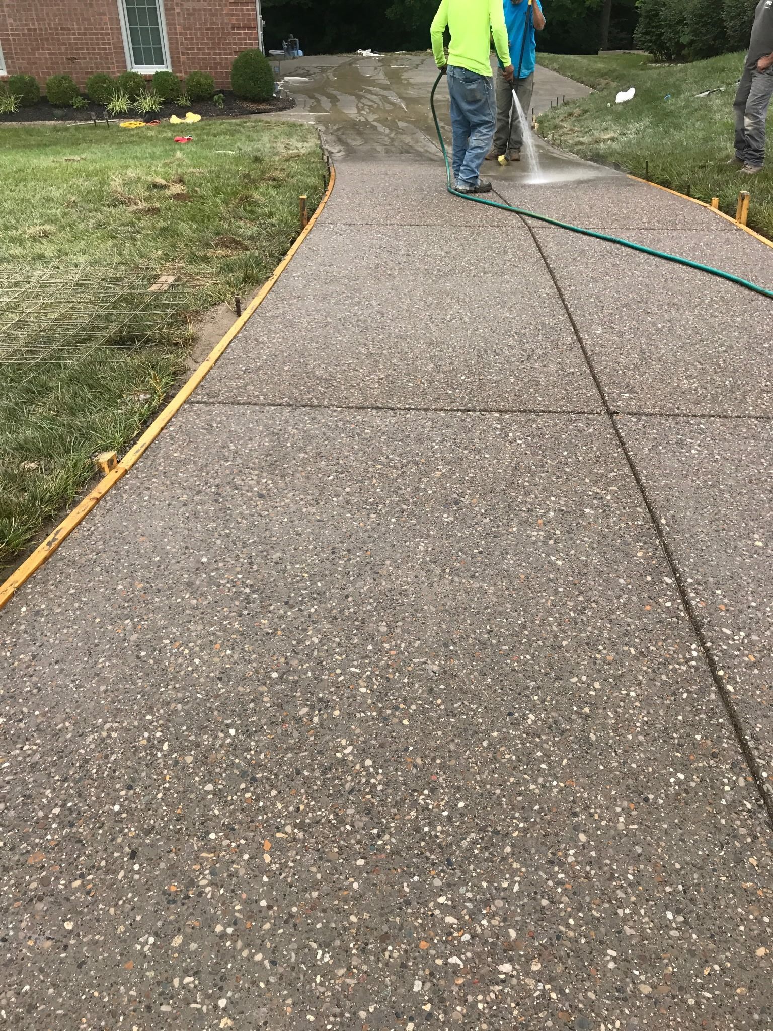 Workers hosing down a concrete driveway, next to grass. Yellow edging is visible.