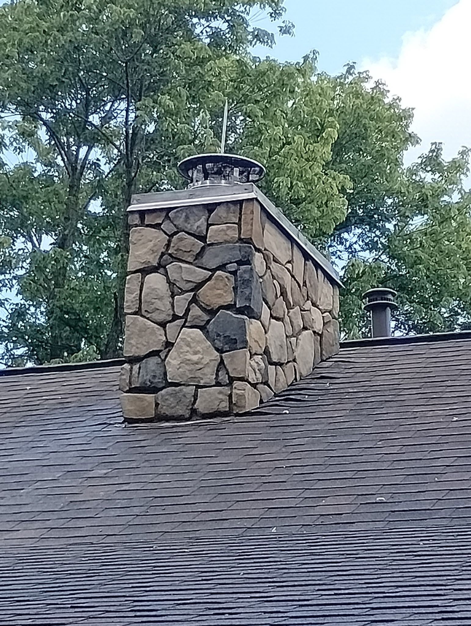 Stone chimney on a dark roof with trees in the background.