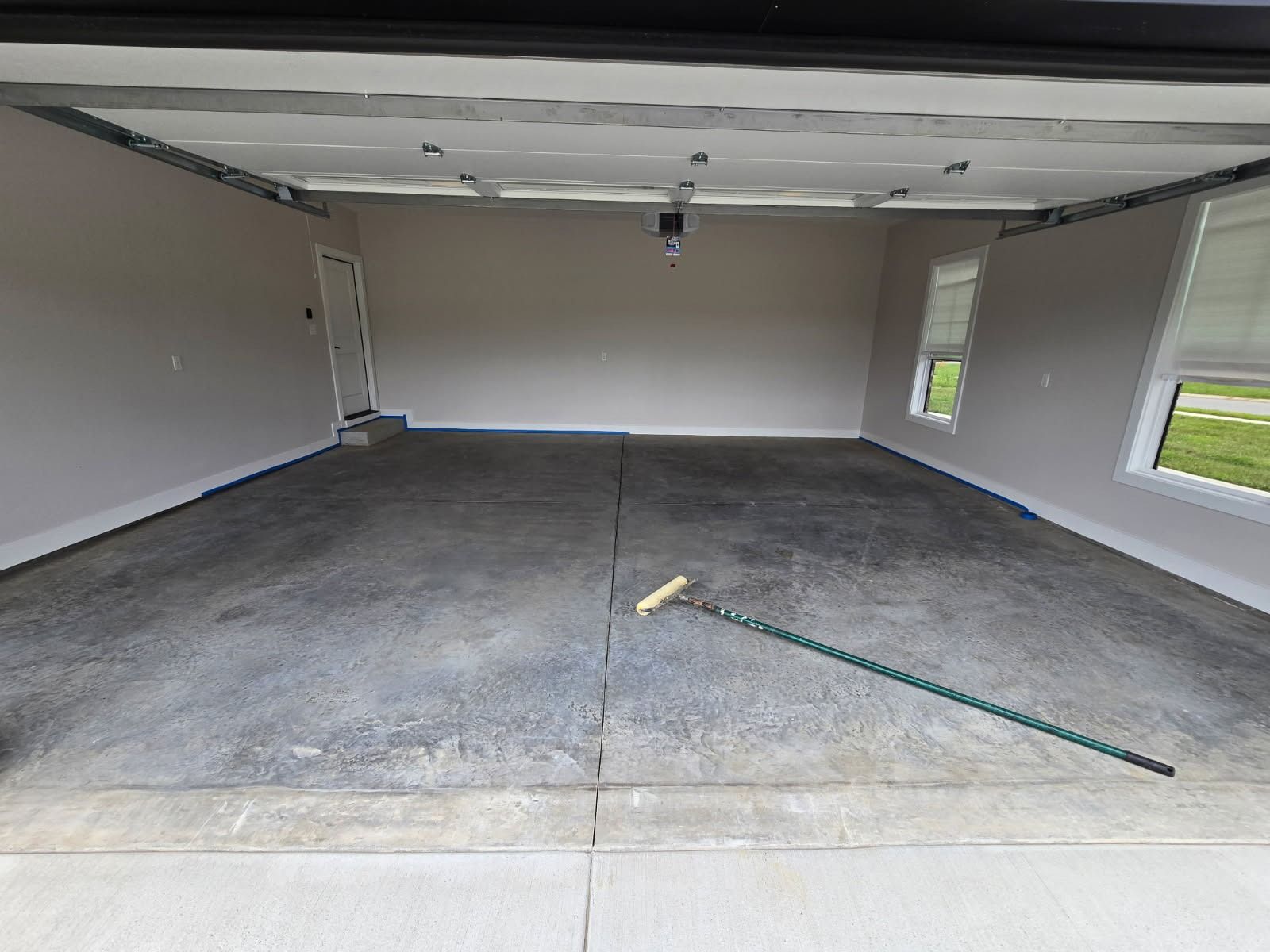 Empty garage with gray concrete floor, beige walls, and a window. A long-handled tool rests on the floor.