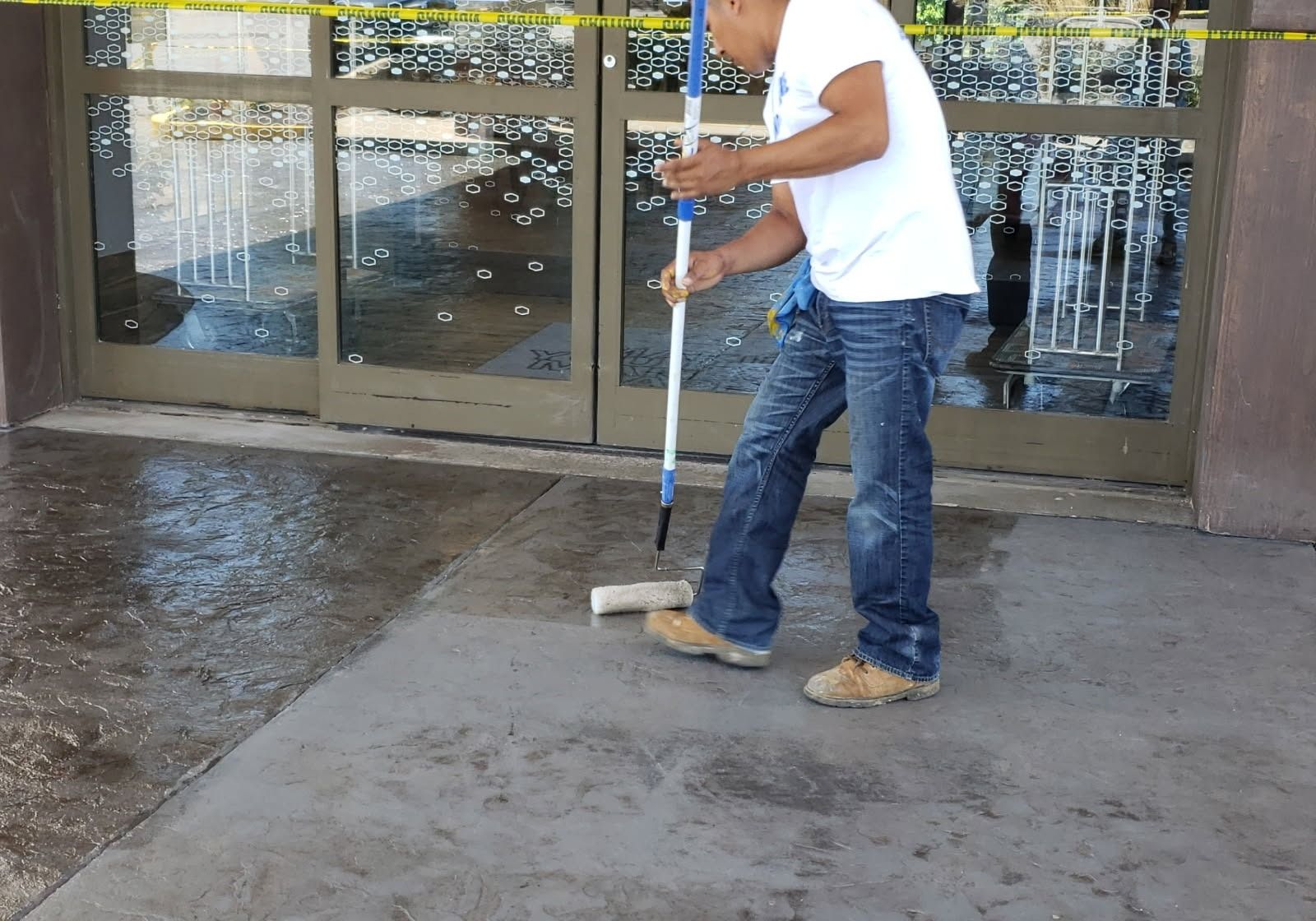 Man mopping wet concrete outside a building with glass doors, yellow caution tape overhead.