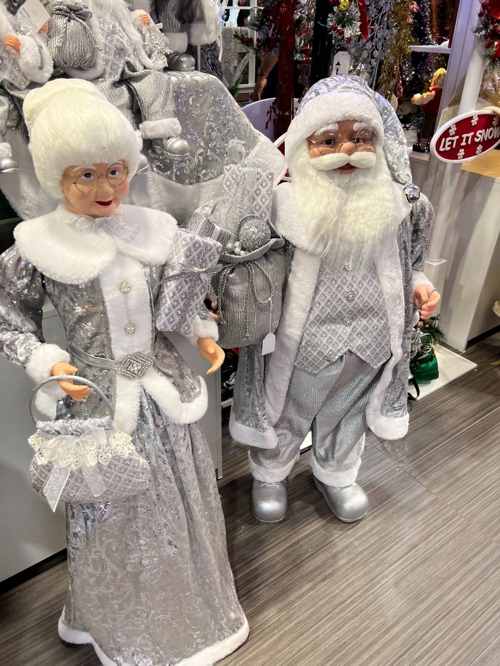 Silver Mrs. Claus holding purse, and Santa in matching outfits, standing indoors near Christmas decorations.