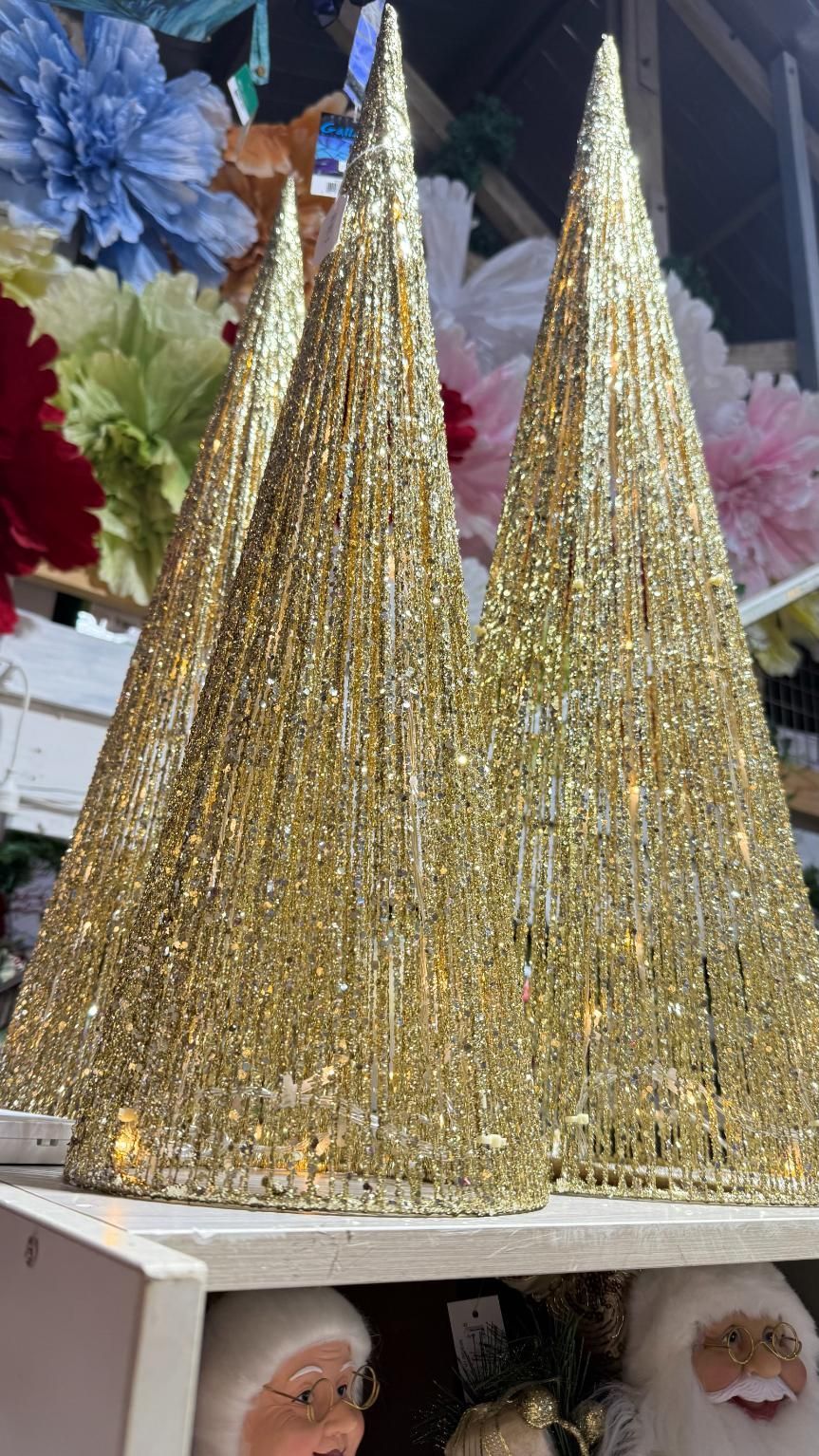 Gold glitter cone-shaped Christmas tree decorations on a shelf, with Santa figures below, and colorful floral decorations in the background. | Jingle Bell Christmas