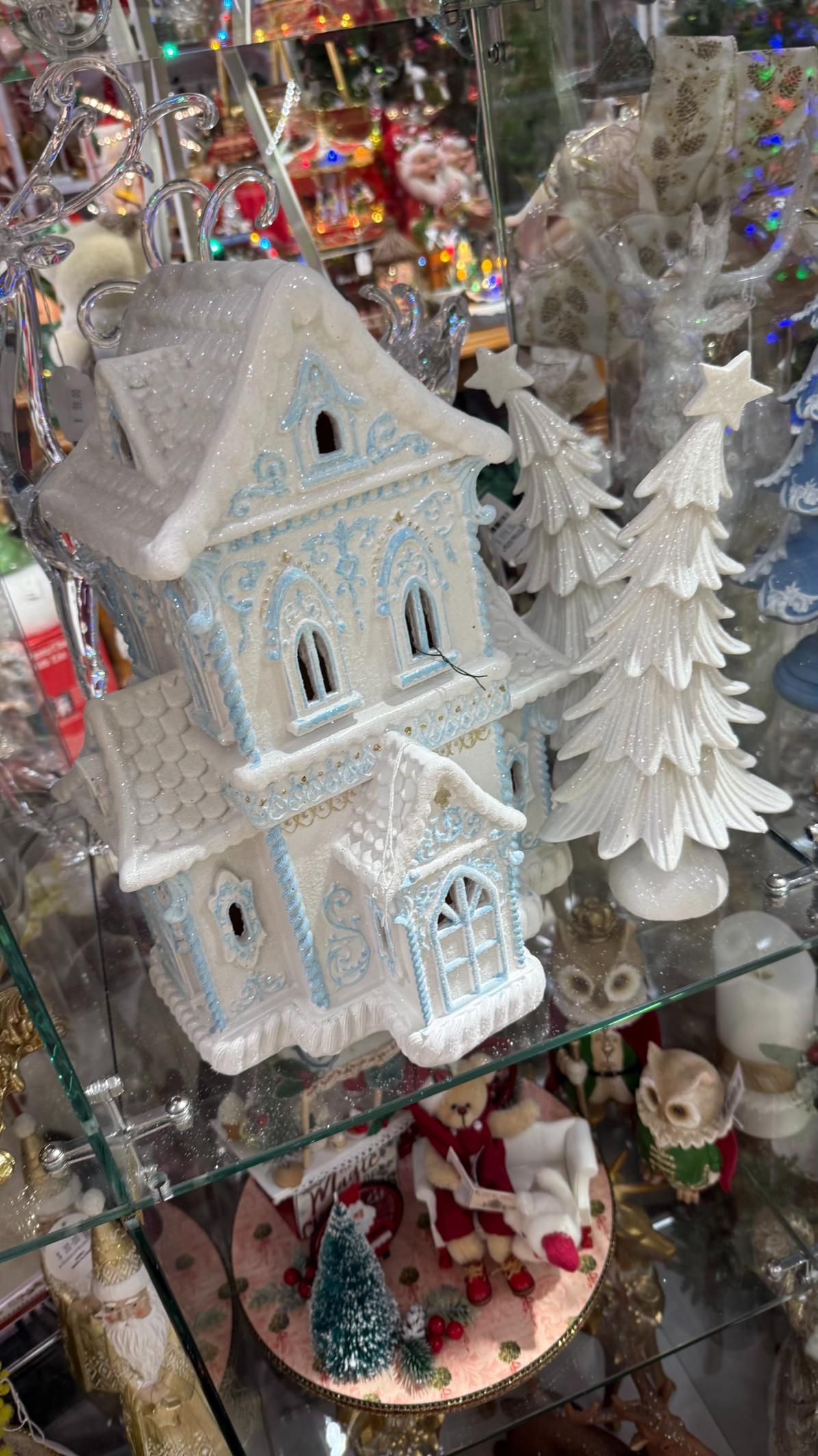 A light-blue and white Christmas house with a Christmas tree sits on a glass shelf among ornaments. | Jingle Bell Christmas