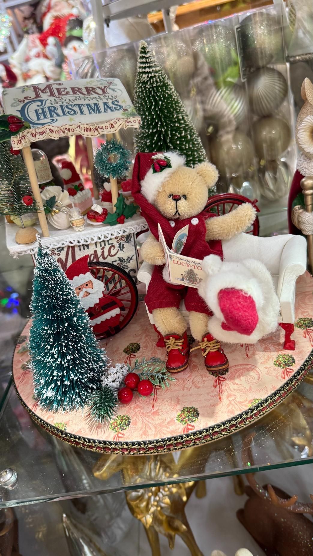 Christmas bear in Santa suit, seated with a bag, next to a sign that says