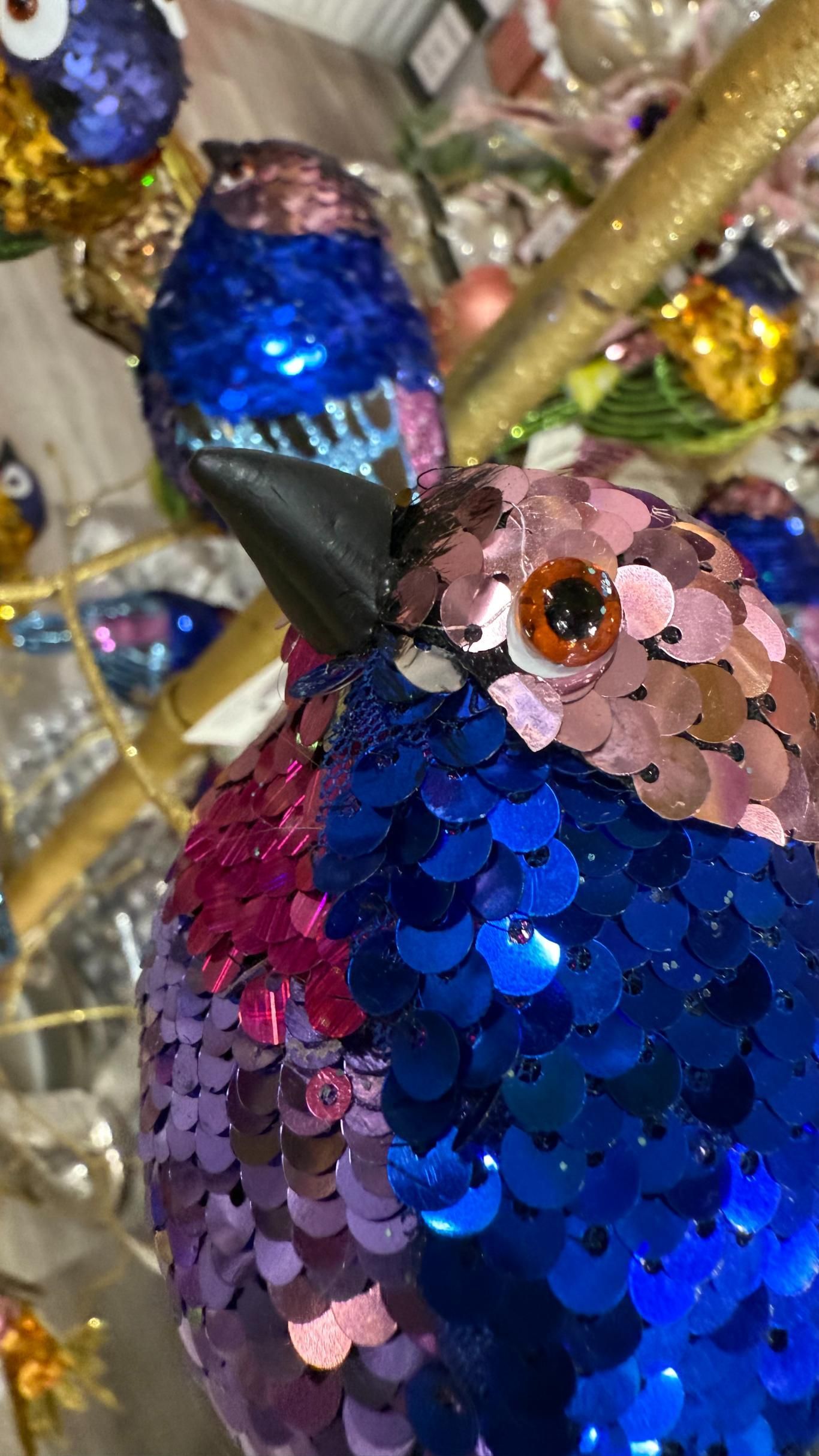 Blue and pink sequined bird ornaments with black beaks and orange eyes, perched on a golden branch. | Jingle Bell Christmas