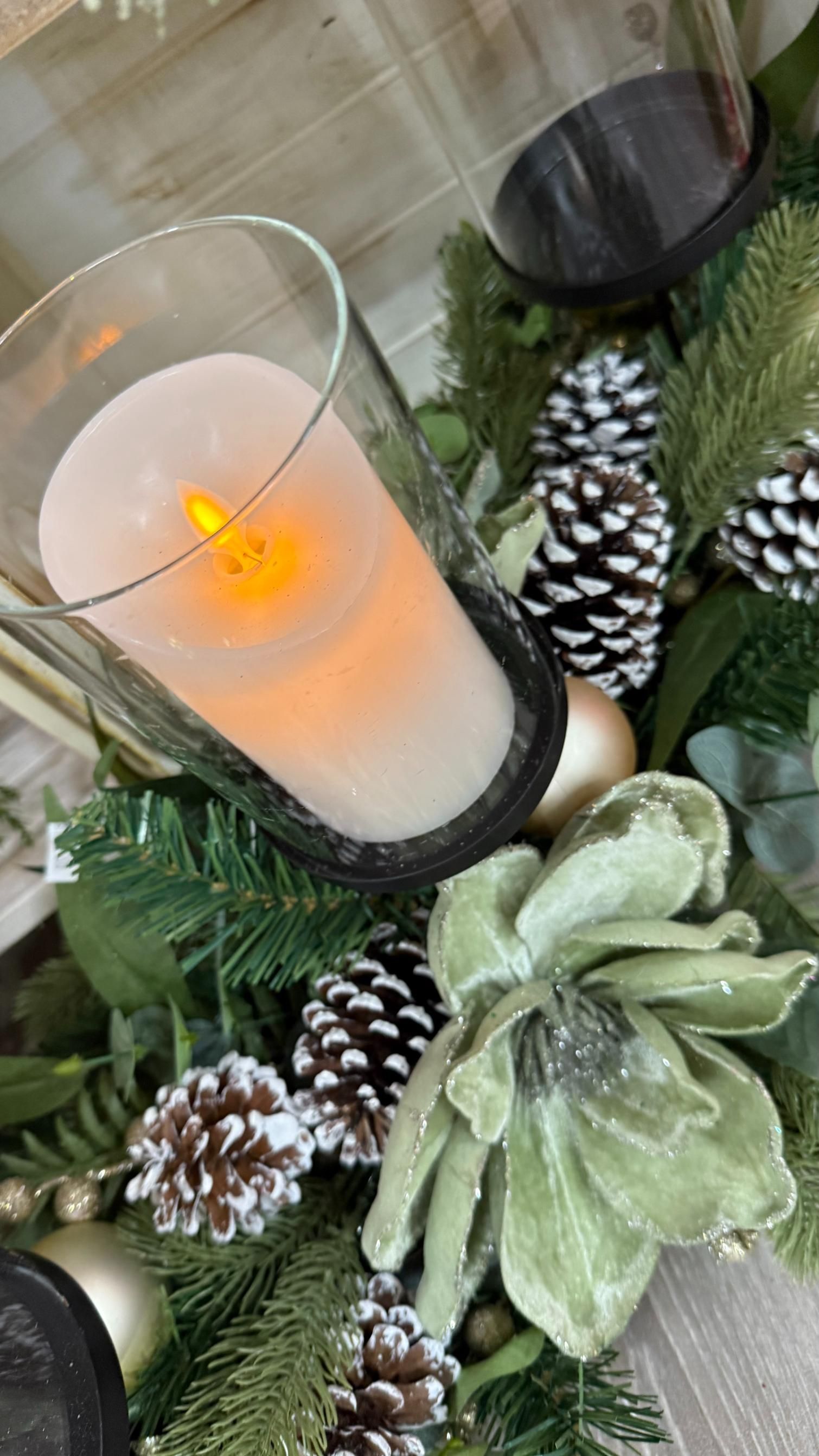 A candle in a glass holder surrounded by pinecones, greenery, and a light-green velvet flower. | Jingle Bell Christmas