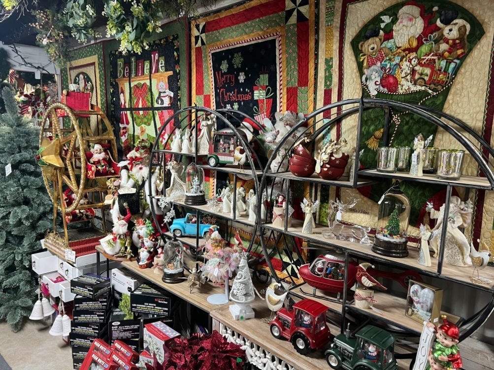Christmas decorations on shelves in a shop, including Santas, cars, and angels, with colorful holiday quilts in the background.
