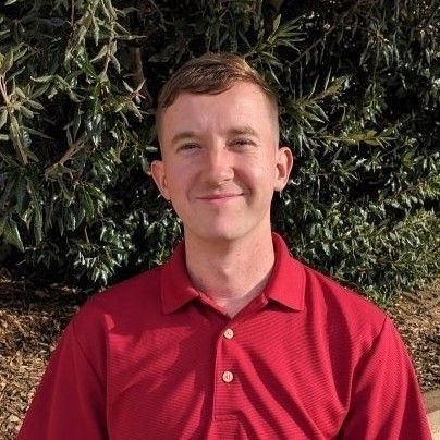 A man in a red shirt is standing in front of a bush.