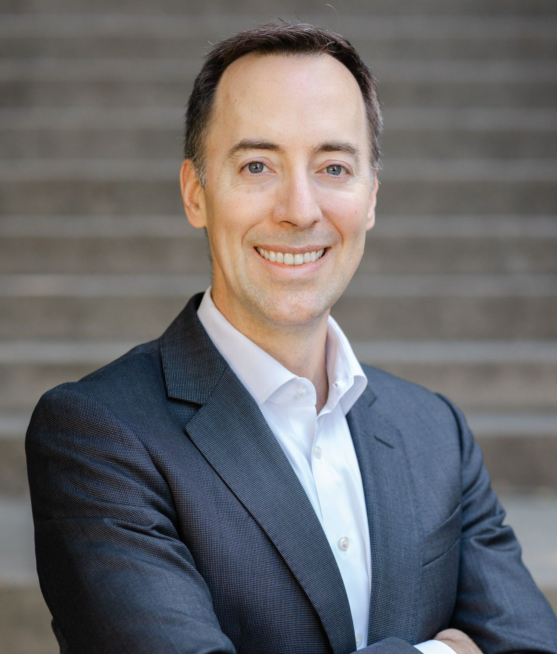 A man in a suit and white shirt is smiling with his arms crossed.