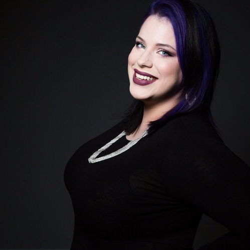 Smiling person with purple hair in a black top against a dark background