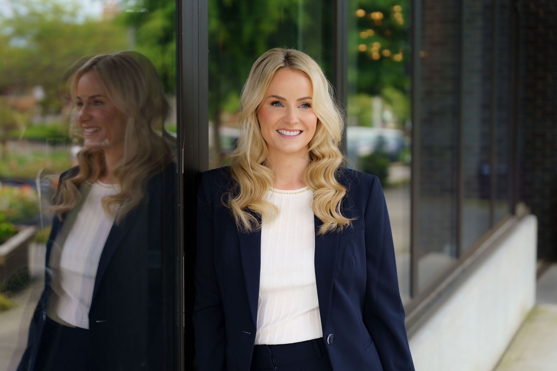 Smiling woman in a navy blazer standing by a glass wall with her reflection visible