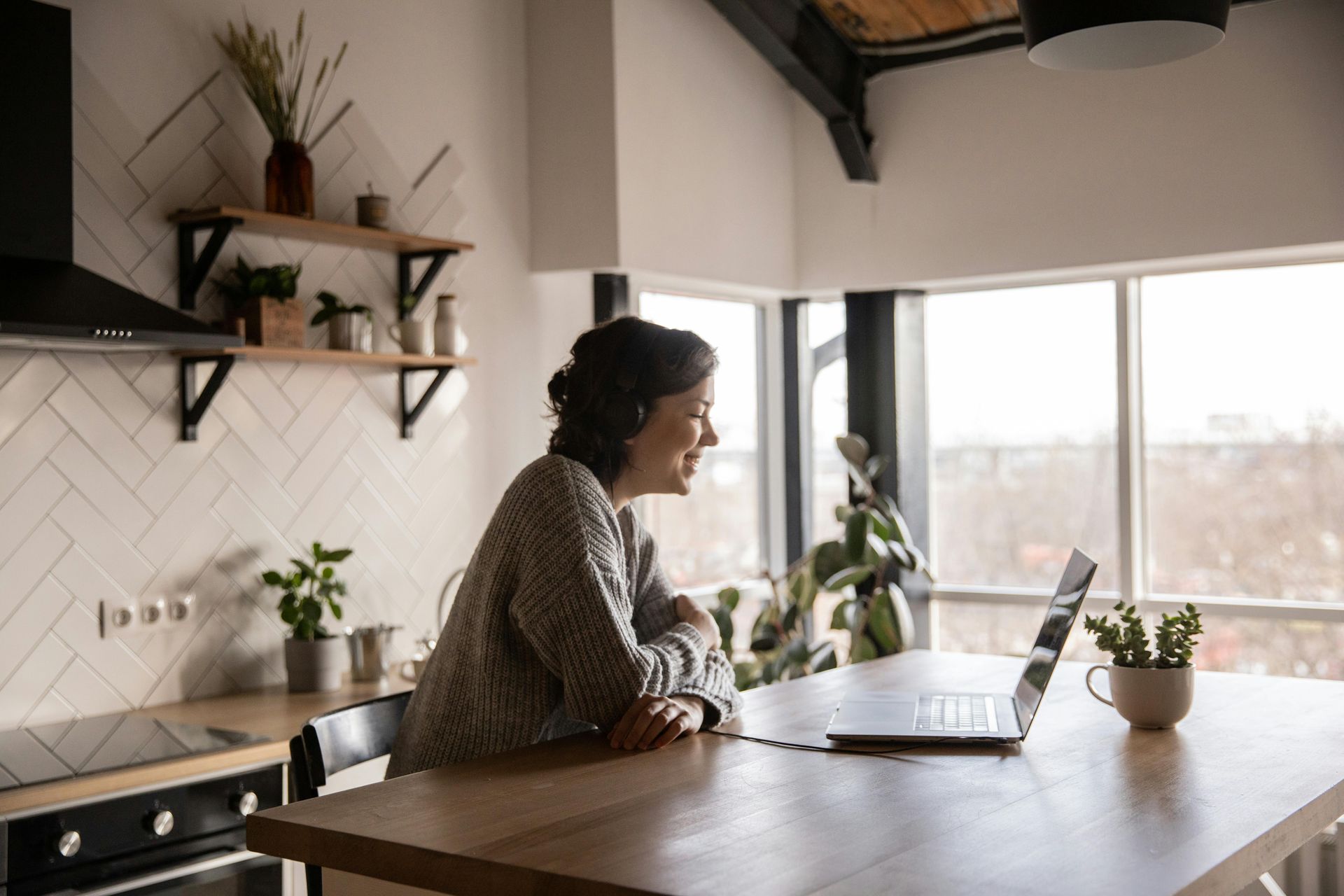 Person working on a laptop at a wooden table in a bright kitchen with plants and shelves nearby