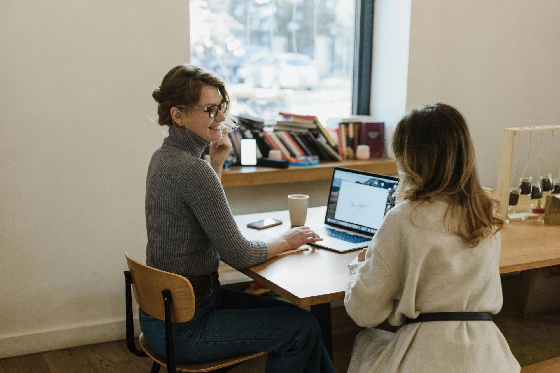 Two people discuss work at a table with a laptop in a bright office