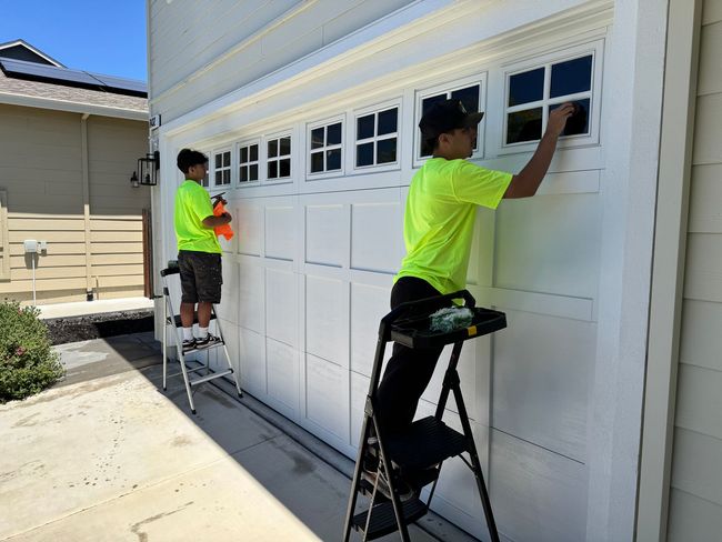 Two workers in neon shirts paint a white garage door and trim outside a house.