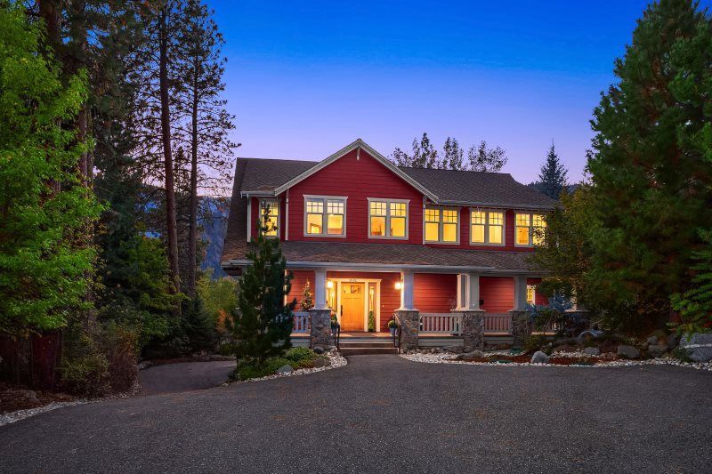 Large lit house at dusk surrounded by tall trees, with a driveway and porch lights on