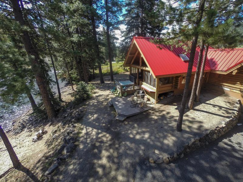 Wooden cabin with a bright red roof among tall pine trees, with a stone patio in the foreground.