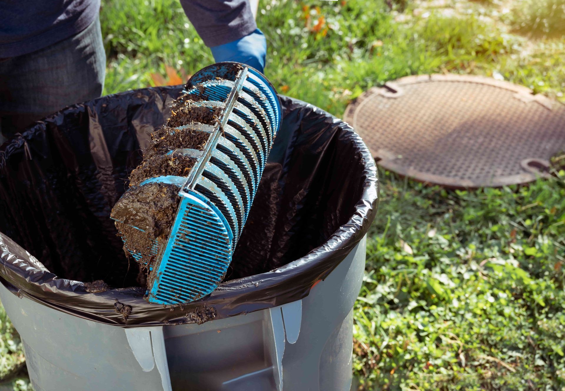 Person emptying a debris filter into a trash can near a sewer access point outdoors.