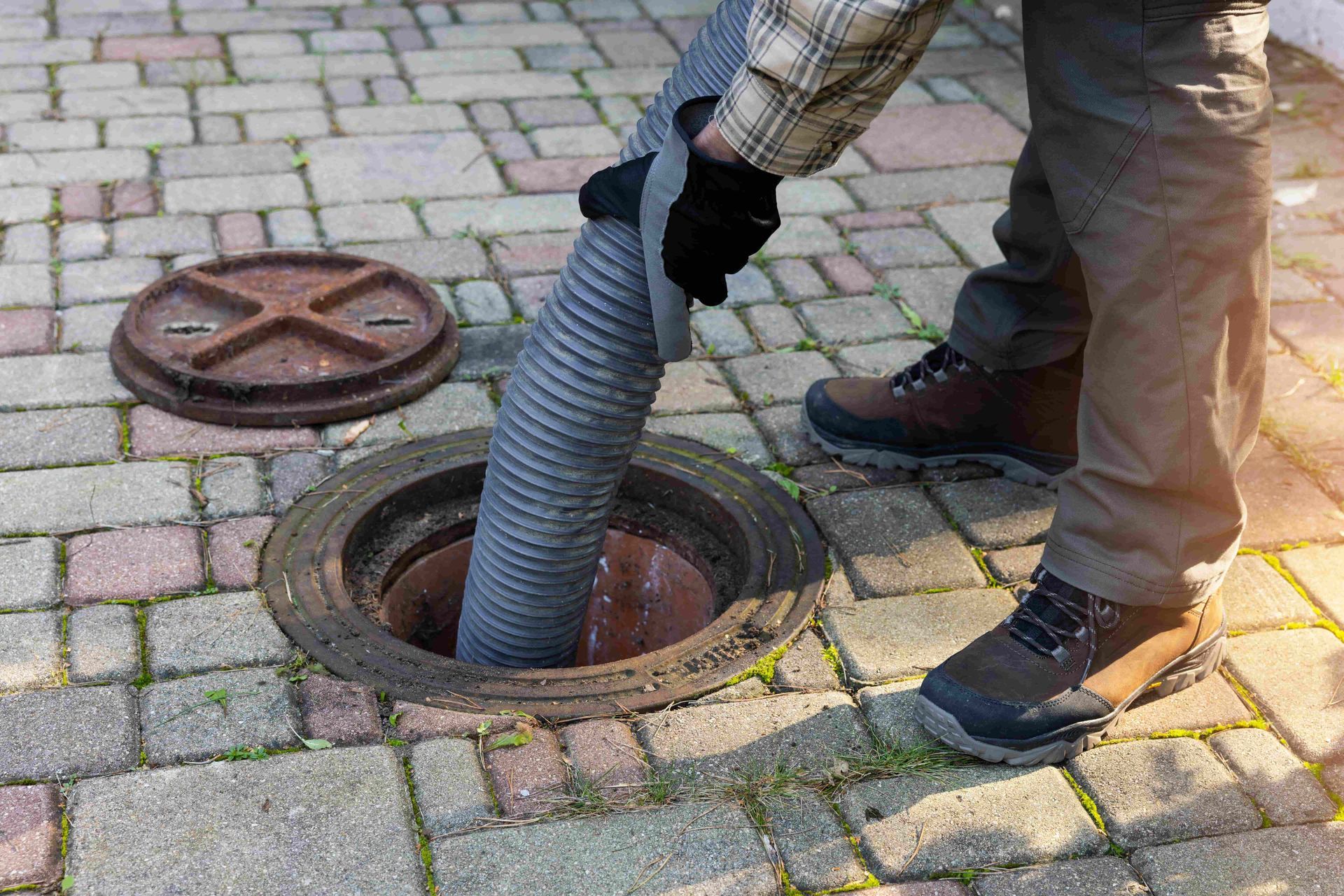 Man in work boots pumping a hose into an open sewer access point in brick pavement.