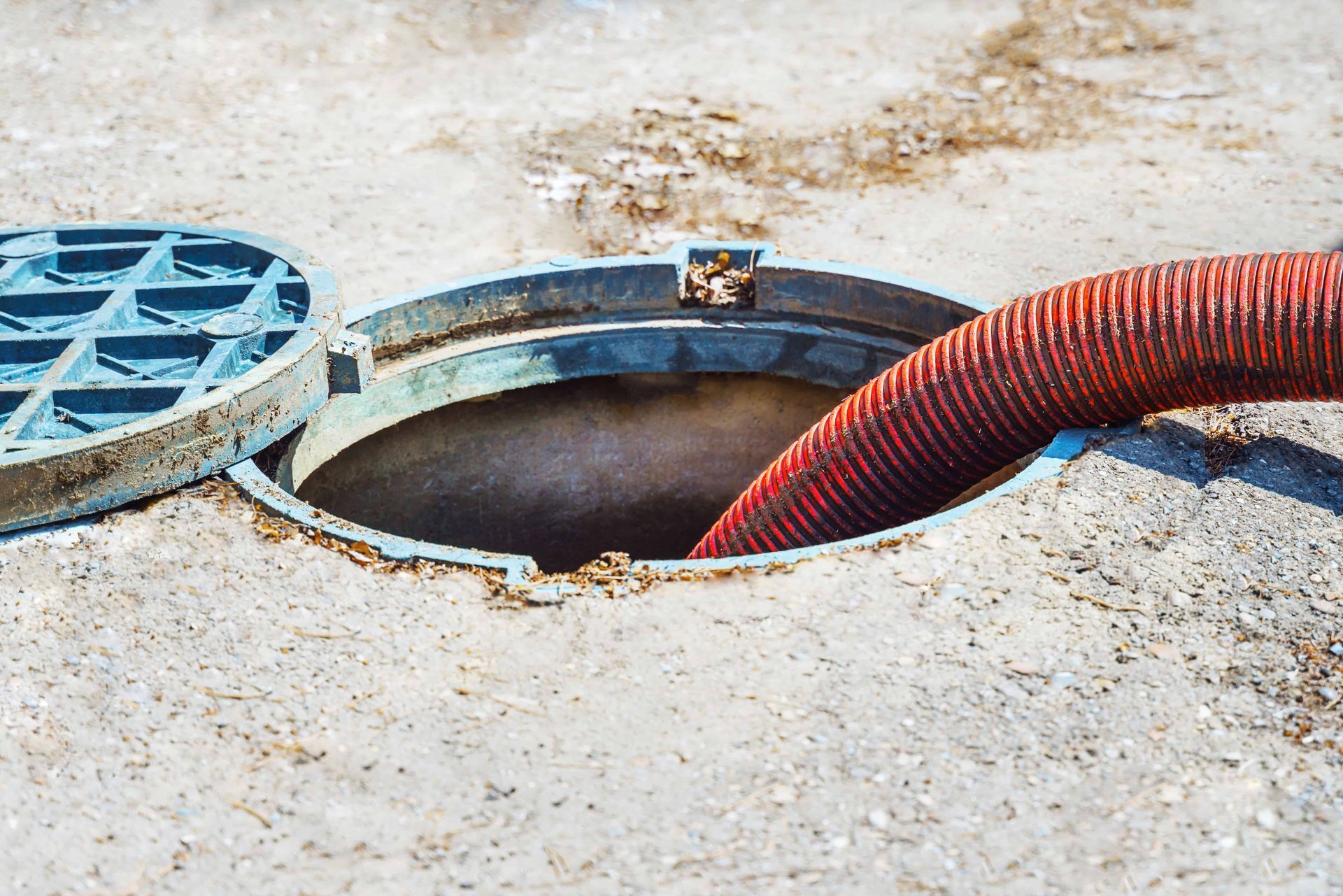 A sewer manhole with the lid open, a red hose inserted, on concrete ground.