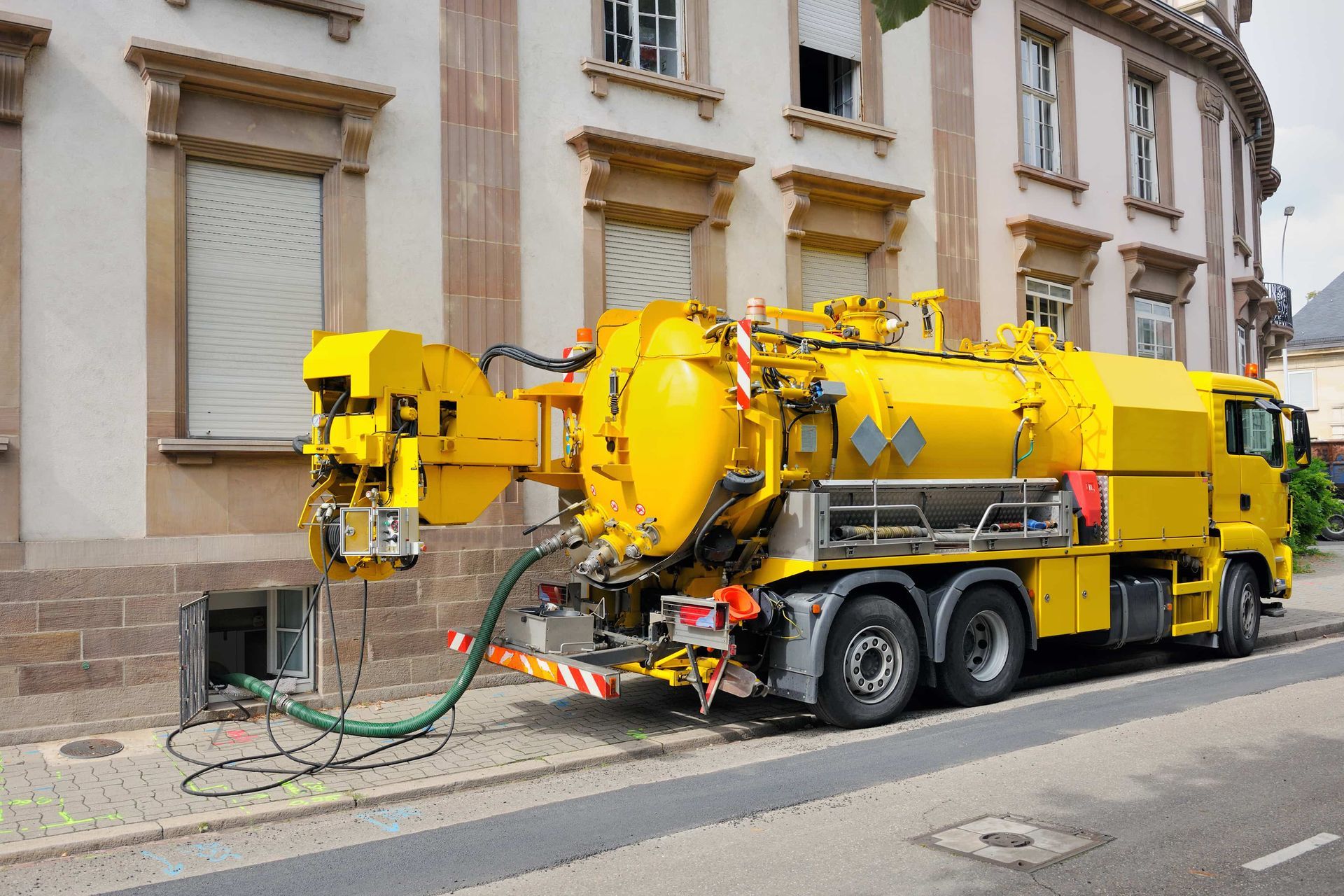 Yellow sewer truck parked outside building, hose connected to underground access point.