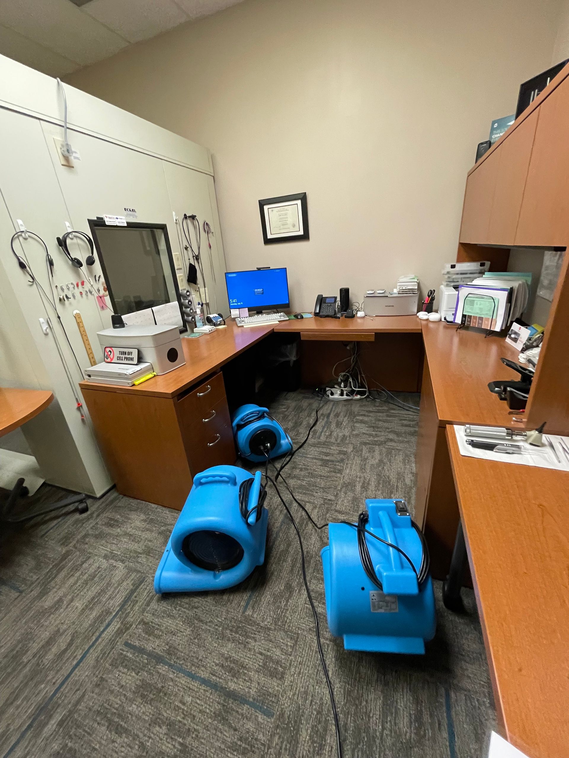 A room with a desk , computer , and two blue dryers on the floor.