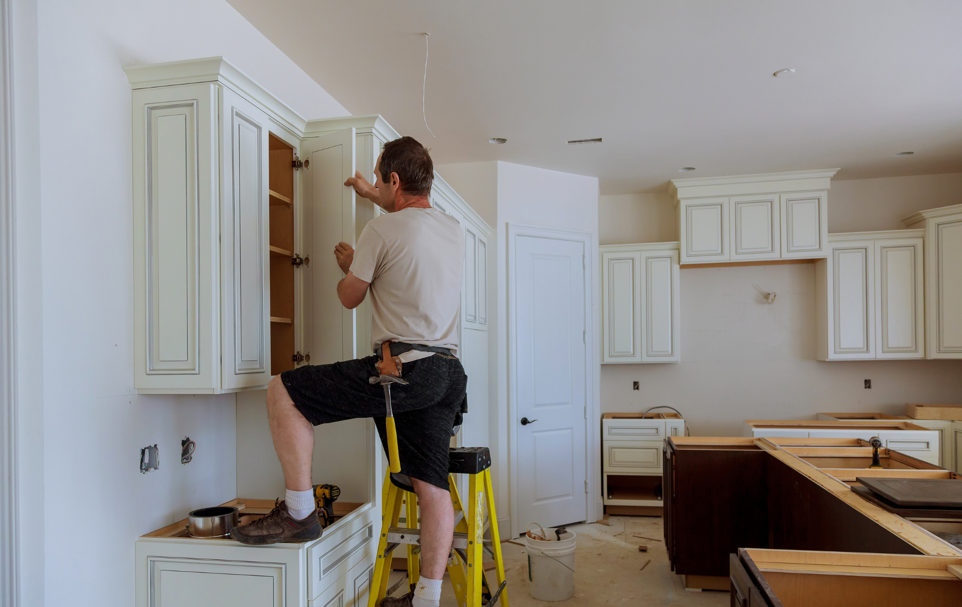 A man is standing on a ladder fixing cabinets in a kitchen.