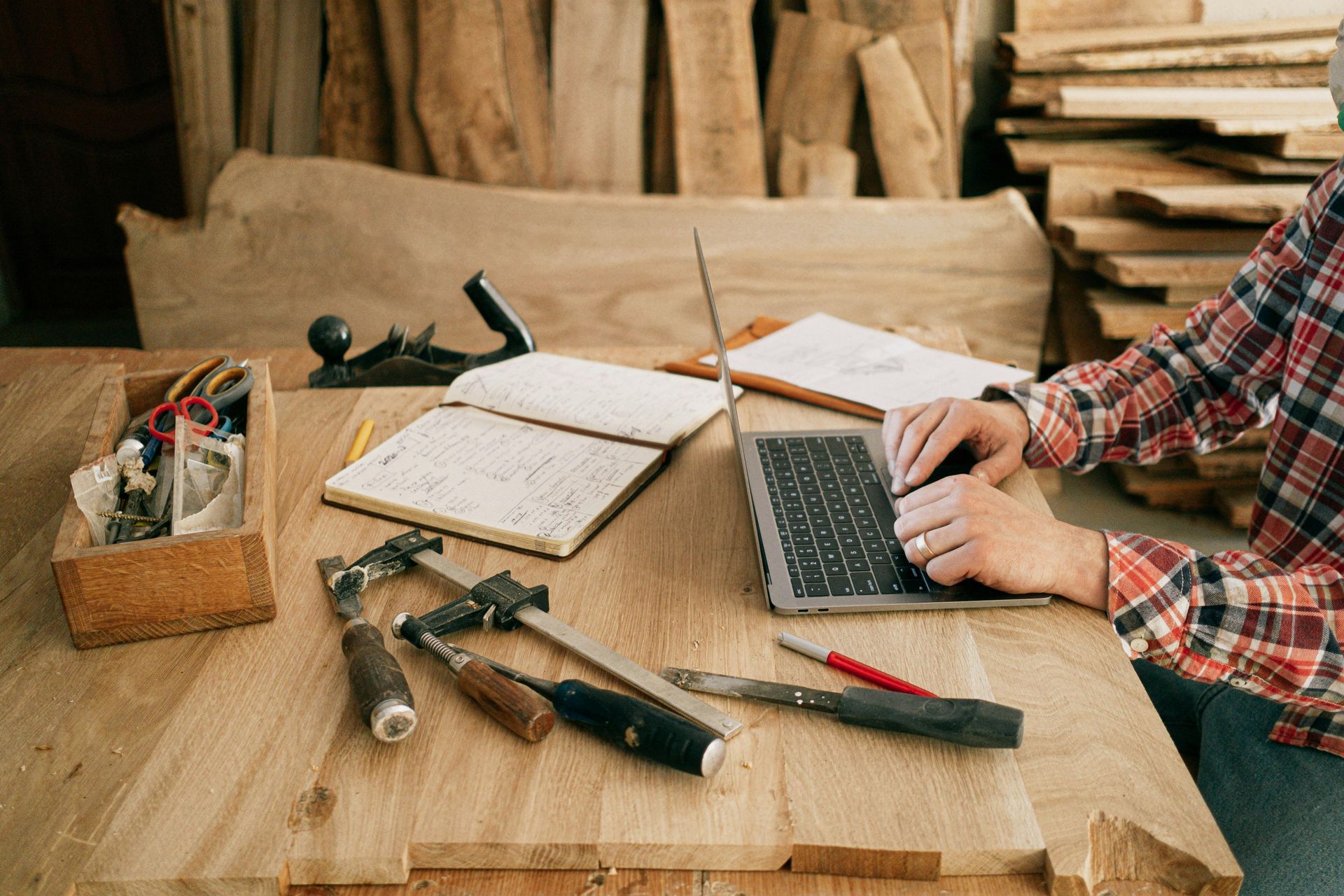 Person using a laptop at a woodworking table with tools and wood planks.