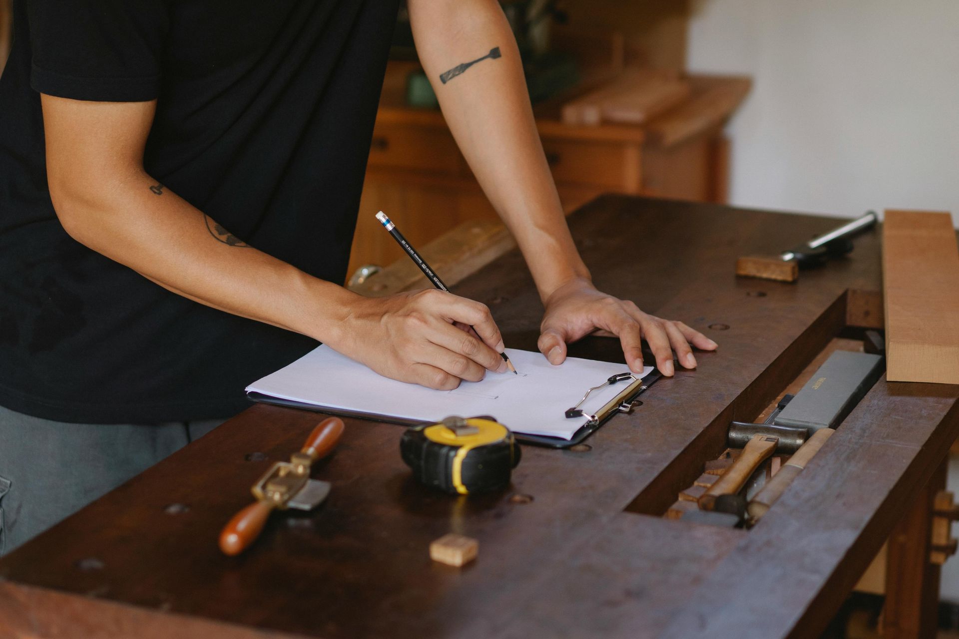 Person sketching on paper at a wooden workbench with tools; close-up of arms and tools.