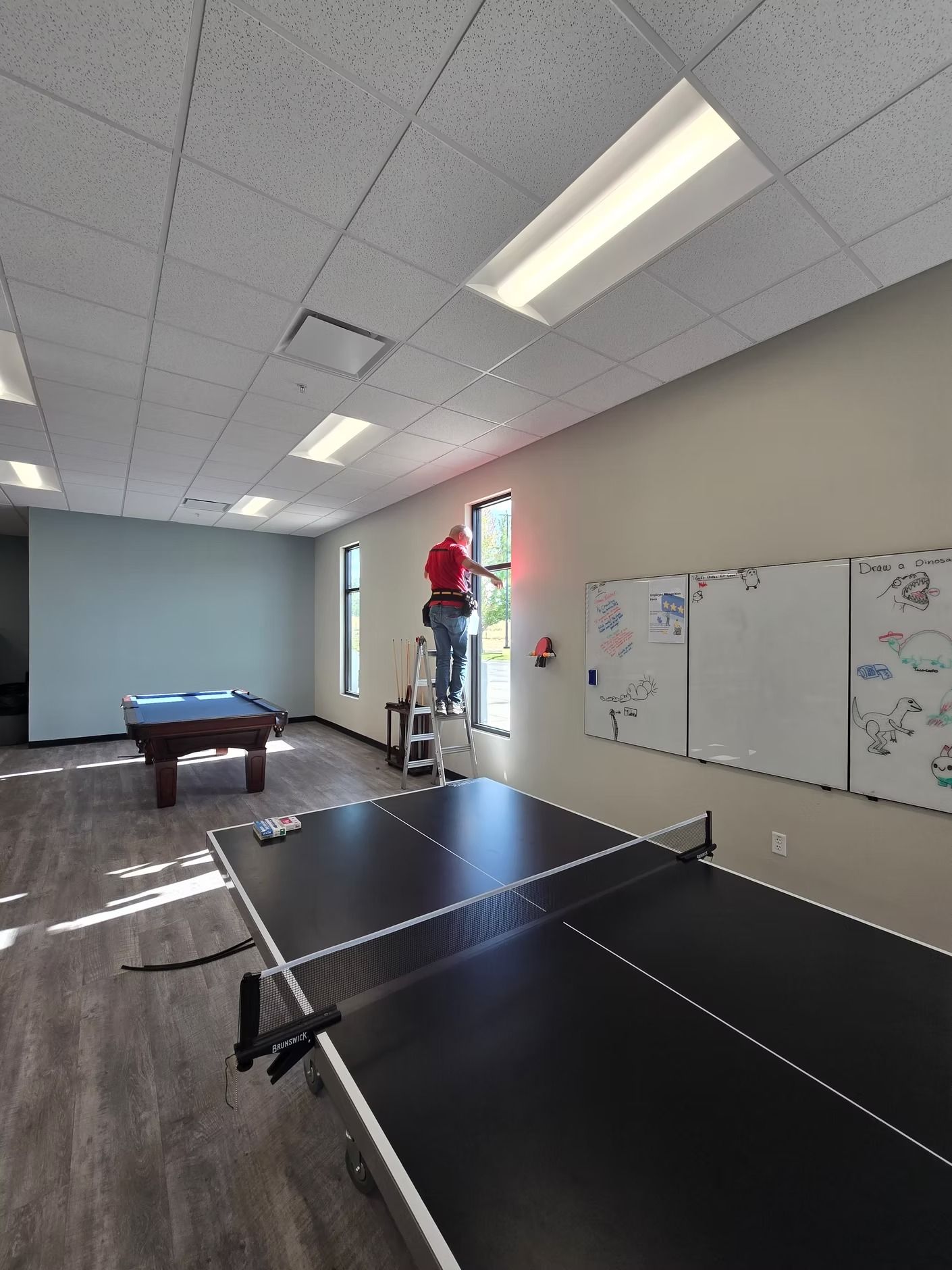 Man on ladder washing window in a room with a pool table, ping pong table, and whiteboards.