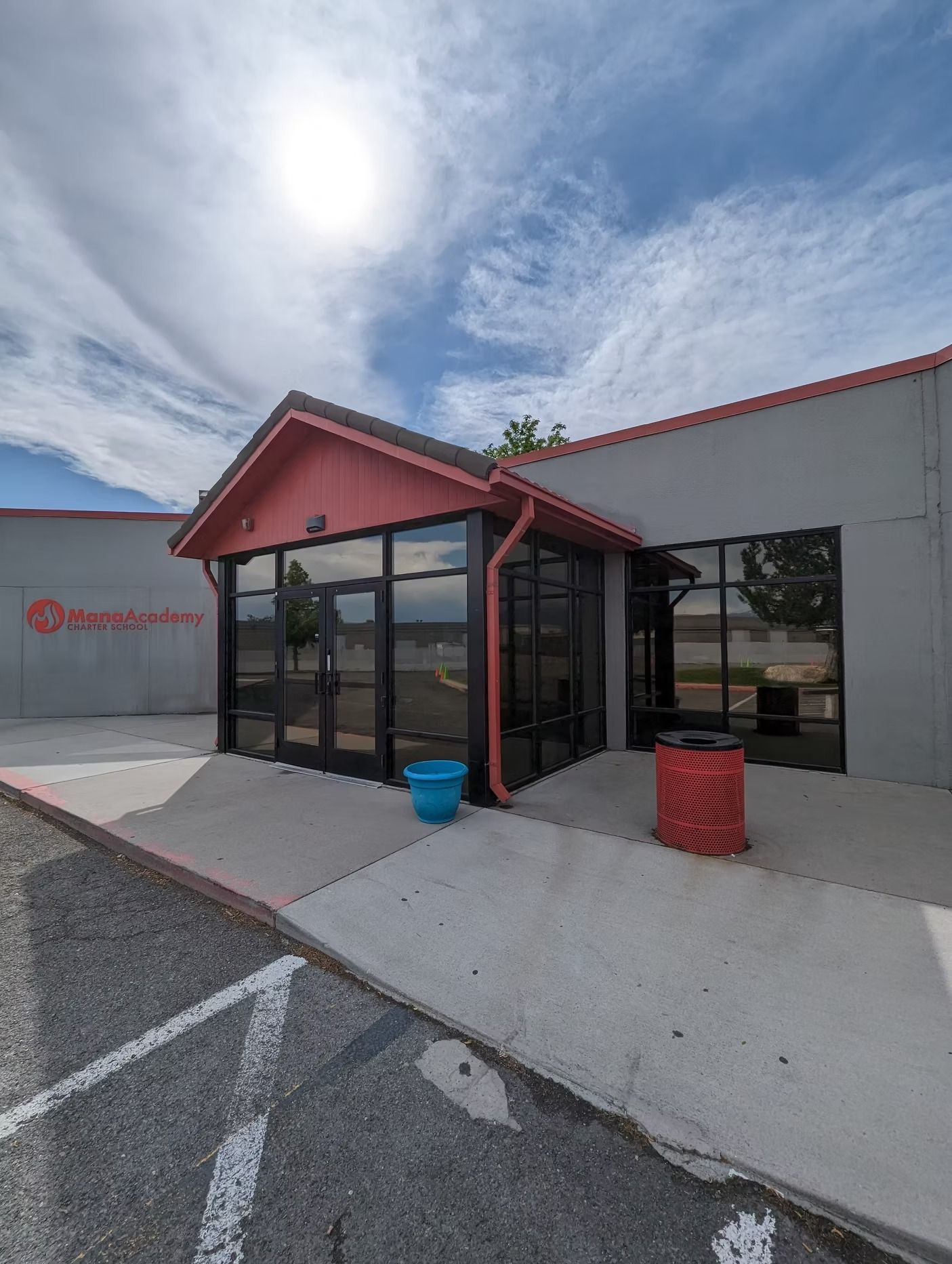 A building with a red-roofed entrance and large windows, a blue bucket, and a red trash can.