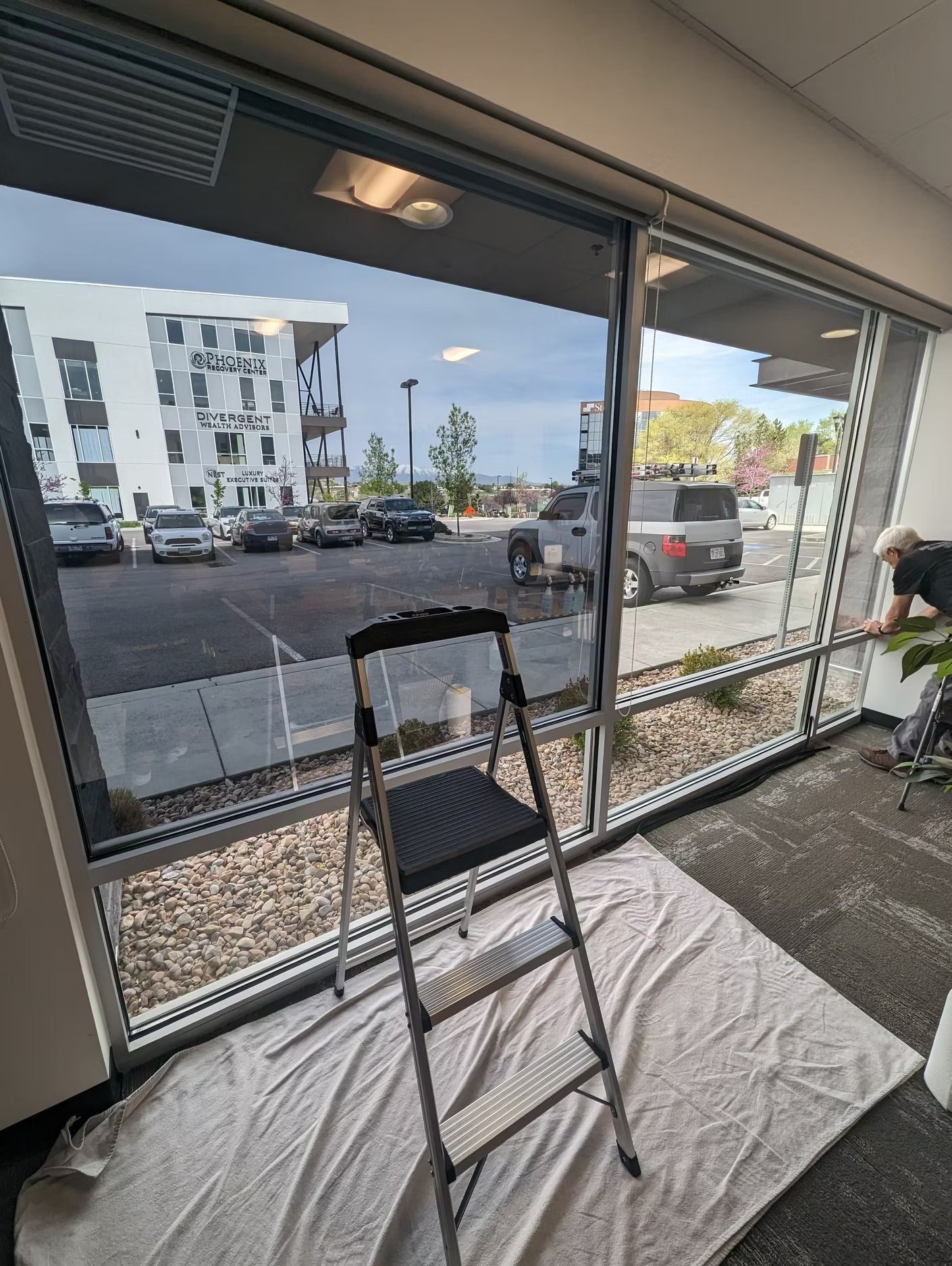A person installing window film in an office. A ladder, window, and outside view of cars and buildings are visible.