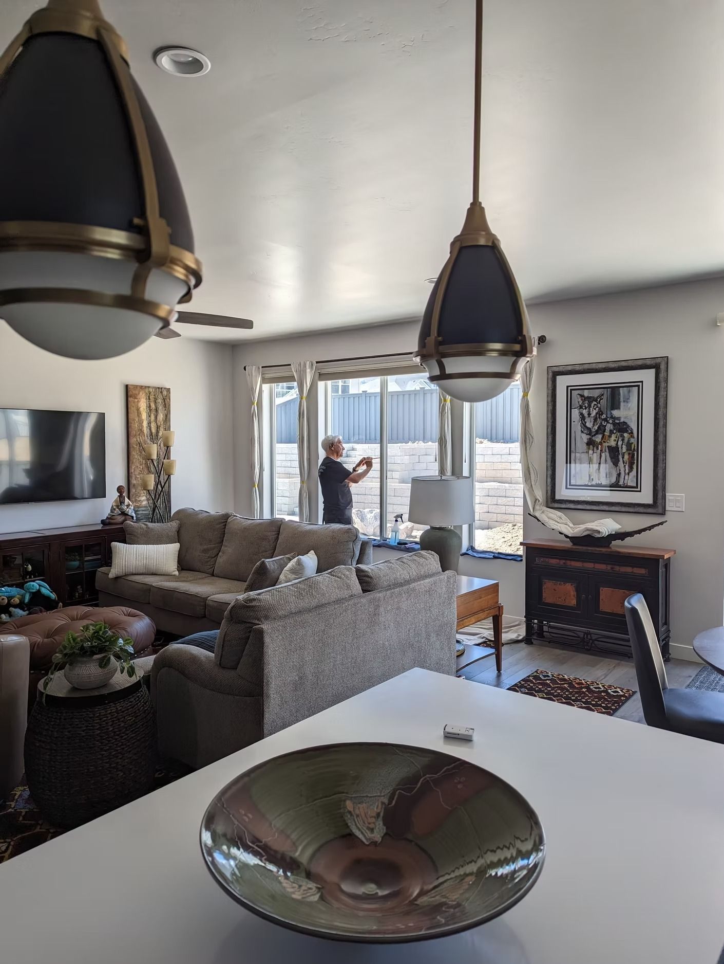 Living room interior with a person near a window, couch, artwork, and a hanging light fixture.