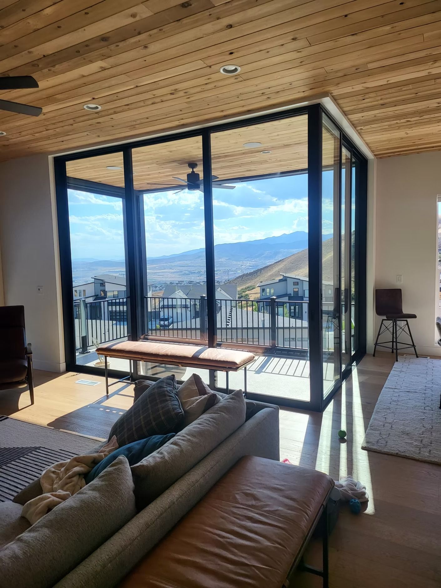 Living room with large windows overlooking a city and mountains. Brown leather sofa, bench, and wood ceiling.