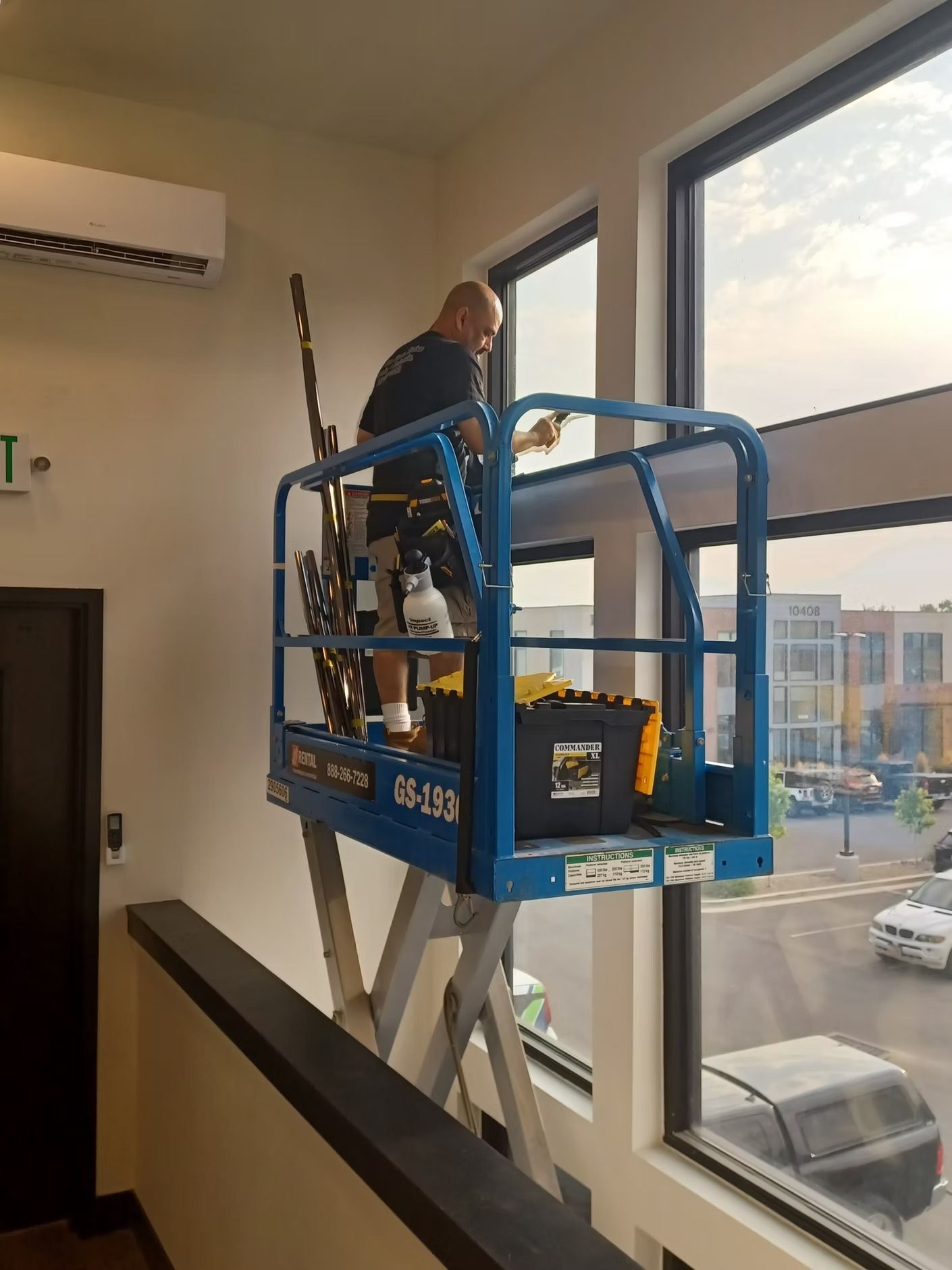 Man on blue lift cleaning a tall window. Exterior view, gray sky, white walls.