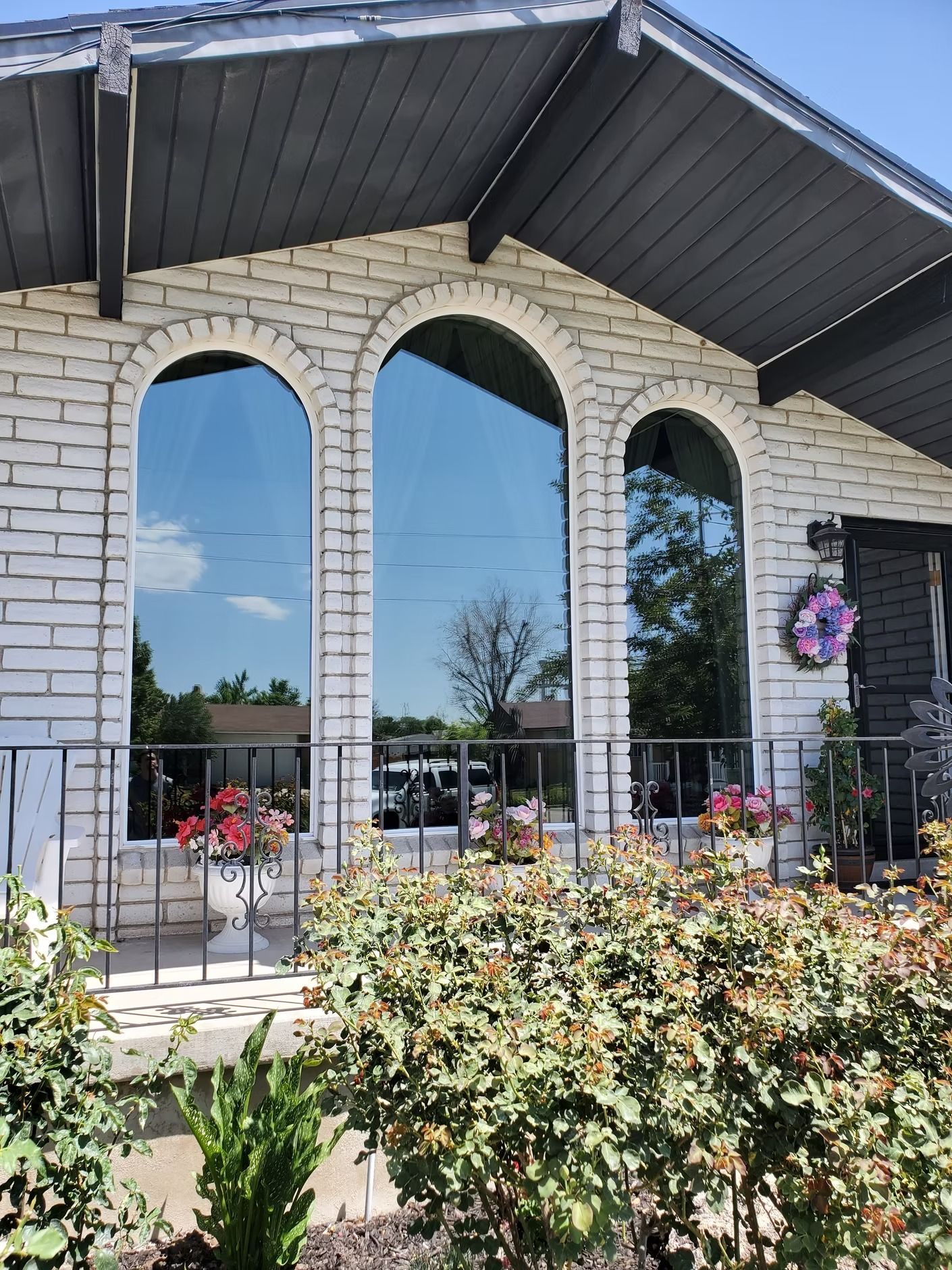 White brick house with arched windows reflecting the sky and surrounding trees. Flowers line the railing.