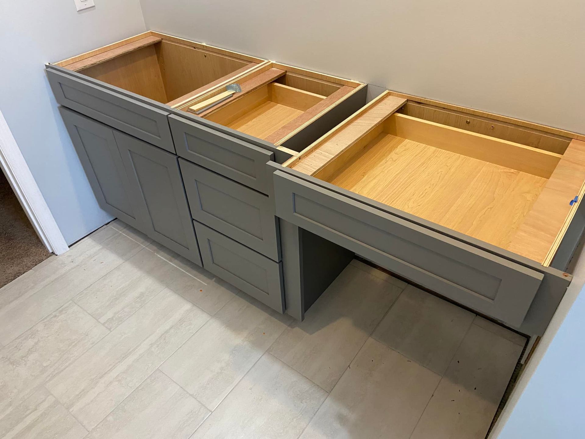 Gray bathroom vanity cabinets installed against a wall with a light-colored floor. Drawers are open.