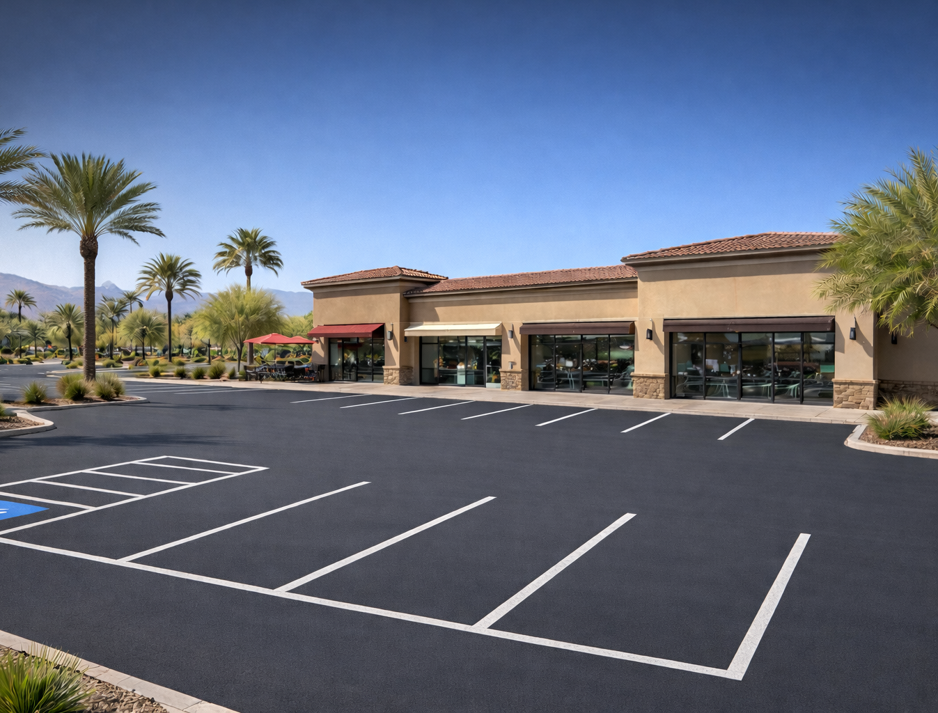 Empty parking lot in front of a tan commercial building with large windows and palm trees under a blue sky.