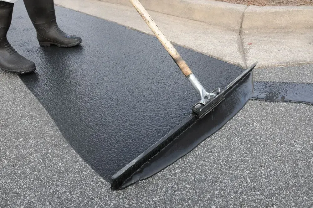 Person in boots smoothing asphalt with a long-handled squeegee on a paved surface near a curb.